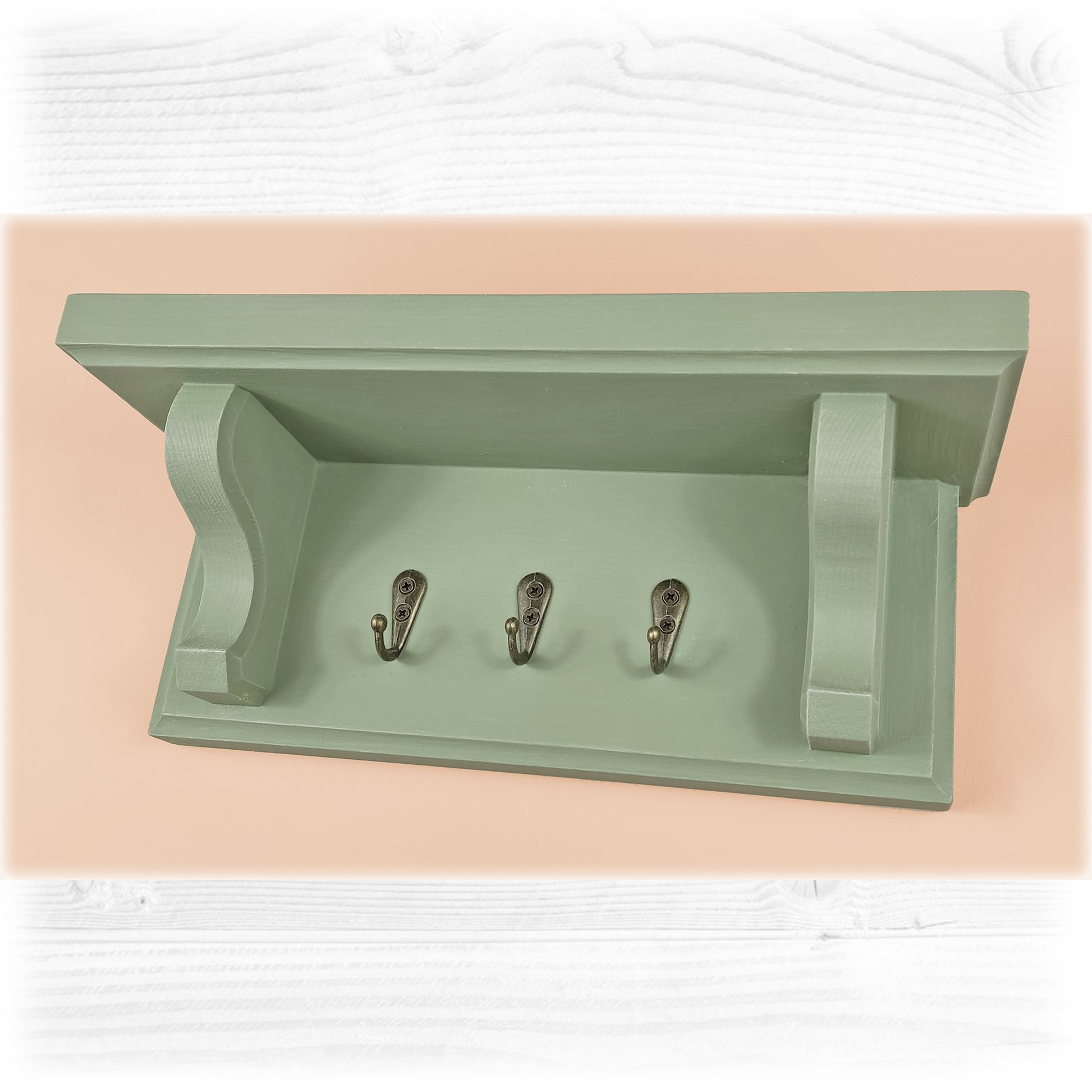 A sage green wooden floating key rack with shelf on a pink background featuring bronze coloured hooks