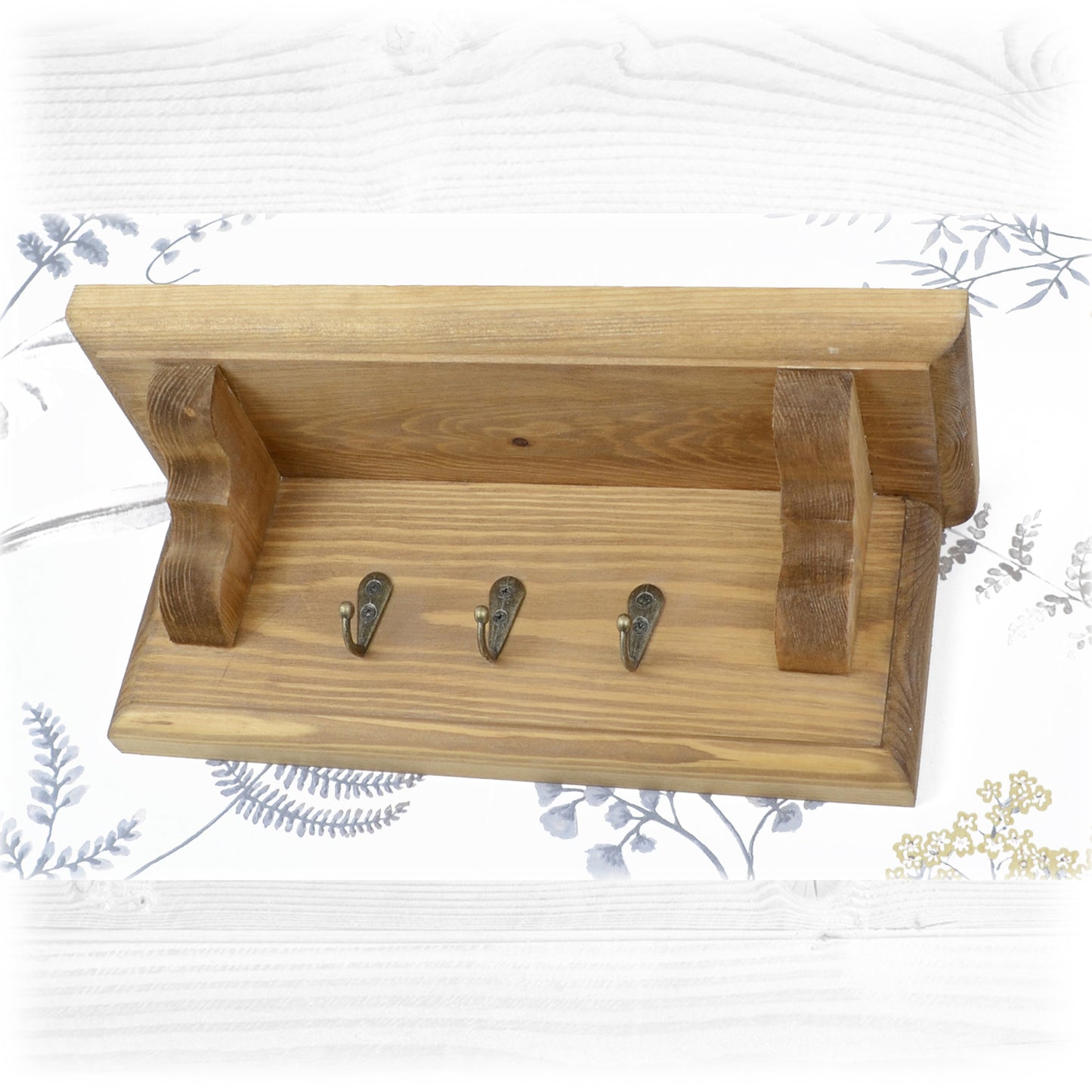 An oak stained wooden key rack with shelf on a floral wallpaper background with bronze coloured key hooks