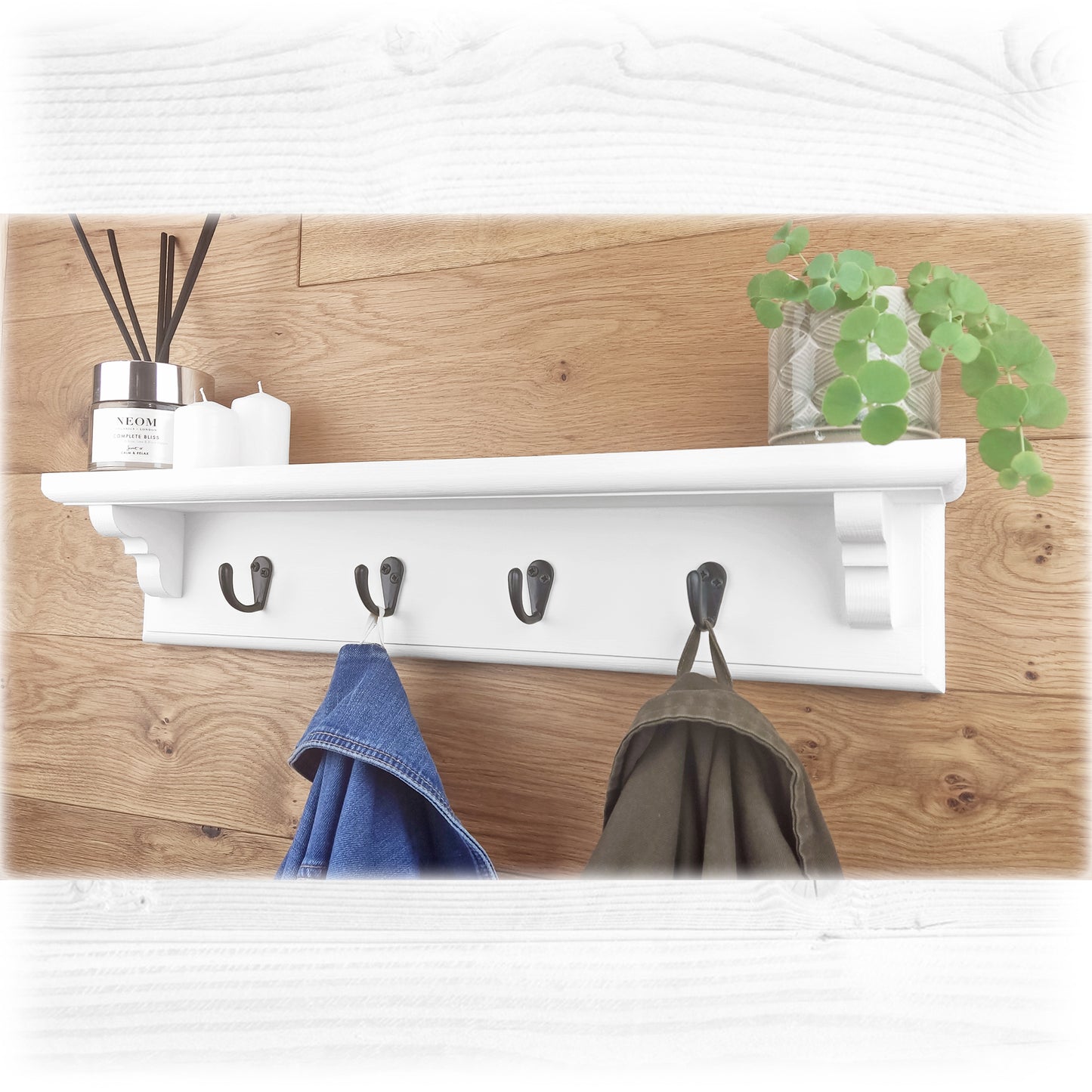 A solid wooden white floating coat rack with black hooks on an oak wooden background holding up 2 coats and with ornaments on top